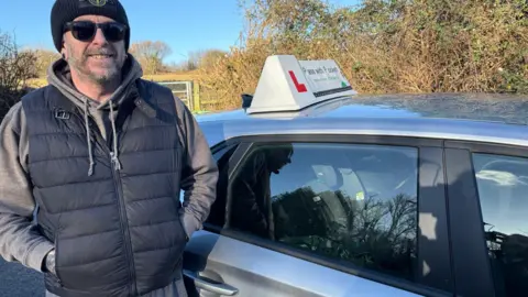 Nathan Pockett stands next to his grey car wearing a body warmer, a woollen hat and sun glasses.