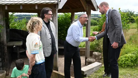 PA Media The Prince of Wales meets locals during a visit to the Apricot Centre in Dartington, Totnes. He's shaking hands with a man outdoors beside a wooden shelter while others stand nearby in a rural landscape.