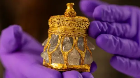 Duncan McLean A rock crystal jar, which resembles an ornate perfume bottle decorated in gold embellishments. A person wearing purple rubber gloves holds the jar.