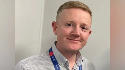 Chris Wilson Head-and-shoulders portrait of a person wearing a light grey button-down shirt and a blue-and-red lanyard, standing indoors against a plain light-coloured wall. The image is well lit and focused on the person's face and upper body.