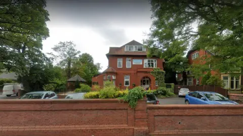 A brick wall with a red brick three storey house and a car park in front