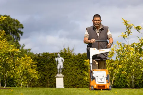 Duncan Mcmillan A man with a moustache and cropped dark hair pushes a lawnmower through a garden