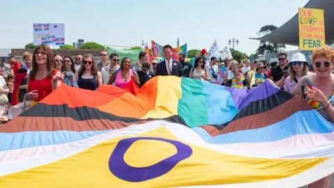 A crowd of people stand behind a Pride flag, which is billowing horizontally, held at the sides by smiling men and women