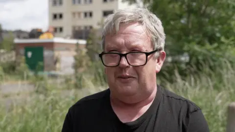 A man wearing a black T-shirt and wearing black-rimmed glasses is looking directly into a camera. He has short grey hair and behind him is a tower block.
