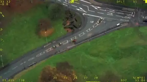 An overhead image of a road junction with a small vehicle, the quad bike, visible in the centre. The junction is surrounded by green land with some trees.