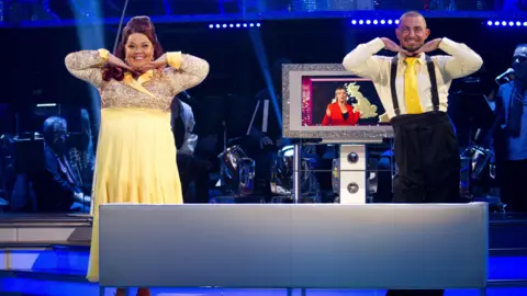 Woman in pale yellow and gold dress with man wearing baggy trousers and pale yellow tie and cream shirt. Both are smiling and have hands perched under their chins.