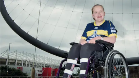 Tom Banks and Paul Thompson Olga is a young girl in a wheelchair with the Riverside Stadium in the background to her left. She is smiling at the camera and wearing a yellow and navy Middlesbrough kit.