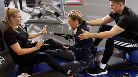 SPINAL RESEARCH Sarra Wilson in the Neurokinex gym in Hemel Hempstead in Hertfordshire where she took part in research funded by the Spinal Research charity. She is sitting on a mat while two physiotherapists - one in front of her, one behind - are helping her use a gym machine.