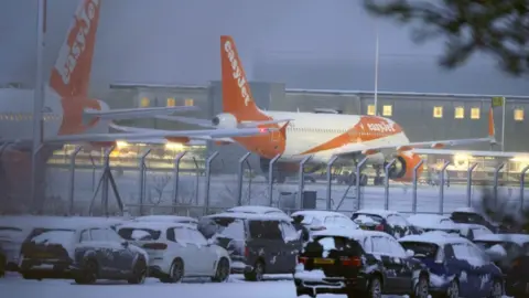 EasyJet planes are grounded due to snow and ice on the runway at Liverpool John Lennon Airport. There are snow-covered cars parked outside the airport's fence.