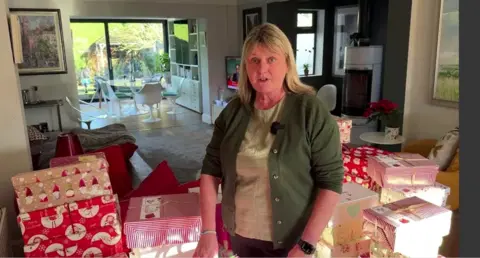 Mrs Rice is stood in her home surrounded by hundreds of shoe boxes wrapped in red and white Christmas wrapping paper