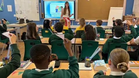 A classroom is pictured from the back with the teacher at the front of class and three rows of pupils sitting with their backs to the camera, some with their hands up.