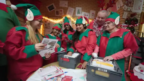 PA Media A group of people dressed as Christmas elves are sorting through boxes of letters sent to Santa. They are smiling at each other as they work. The walls behind them are decorated with children's Christmas cards and light up tinsel. 