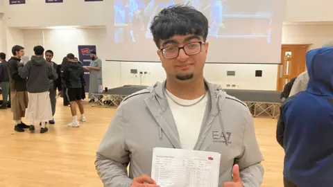 Grace Wood/BBC A teenage boy standing in a large indoor hall holds up a printed results sheet showing school grades, with other people talking near a stage.