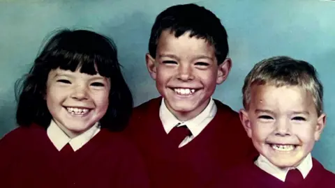 Family photo A photo of Shaun McLaughlin between his younger sister and brother.  They are all wearing red school uniforms and smiling broadly at the camera