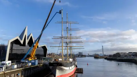 A sailing ship docked next to a large museum building. A large crane and other construction equipment are next to it, with wok clearly taking place on the boat.