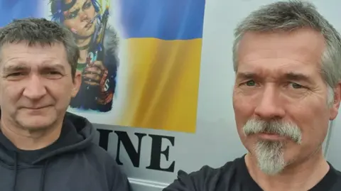 A close-up photo of two men wearing dark tops who are standing in front of a van, which includes artwork on the outside. The man on the left with mainly dark hair and the man on the right with grey hair are looking at the camera.