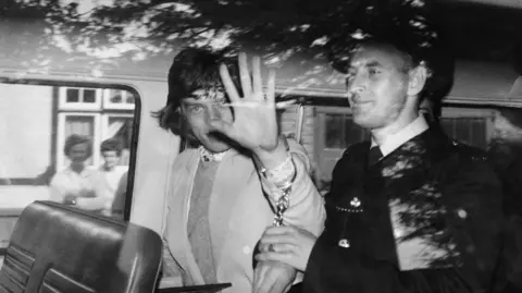 Corbis via Getty Images Police taking away Mick Jagger after Jagger was found guilty of the illegal possession of pep pills in 1967. Mick Jagger is in handcuffs and putting a palm up to the camera