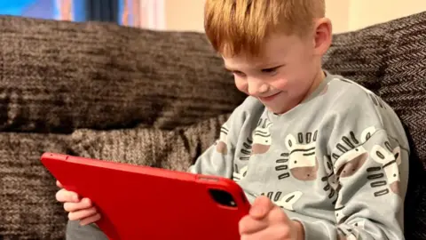 Theo in a zebra jumper, sitting on a brown sofa and reading on a red tablet