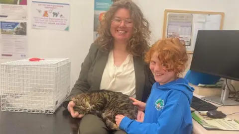 Chloe McCrea A woman sits in a vetinary practice with a tabby cat on her lap. A child stands next to her stroking the cat.