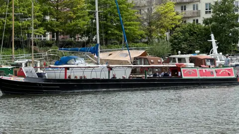 A green and red narrowboat on a city harbour. Passengers can be seen on the foredeck. A Row of moored boats is visible behind. 