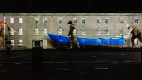 A projection on the barracks of men pushing a blue fishing boat. the three-storey barracks are made of grey stone and have ornamented windows with white panes. 