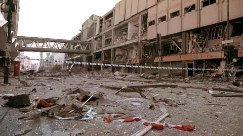 PA Media Scene of devastation following the IRA bomb in Manchester on 15 June 1996. Rubble litters the street and there is a row of blue-and-white police tape in the background, as well as a bridge across the road.