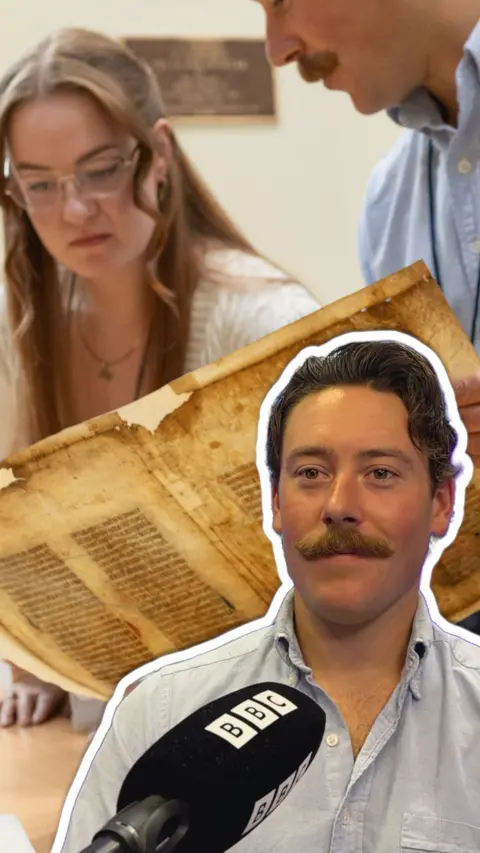 A man with brown hair and a moustache wearing a white shirt in front of a man and woman reading a yellow strained manuscript