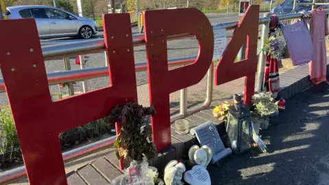 Big red "HP4" letters affixed to crosswalk railings representing Harry Parker and his football jersey number, along with other football jerseys, flowers, candles and other tributes that make up a memorial to the Akers Way teenager.