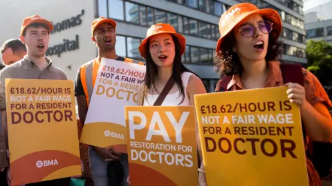 Getty Images Four doctors connected strike, wearing orangish BMA bucket hats and holding up placards calling for much pay, saying that '£18.62 per hr is not a adjacent costs for a resident doctor'. In nan inheritance St Thomas' Hospital is visible. Taken successful London successful July.