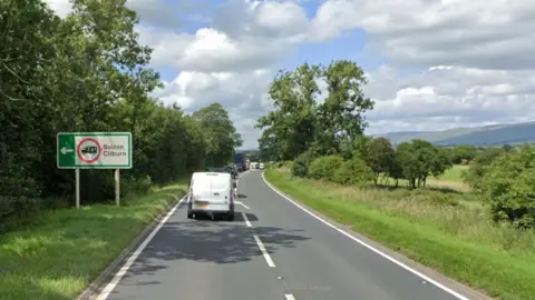 Queuing traffic on a section of road, with green, leafy verges. There is a motoring sign with a left-pointing arrow, saying Bolton and Cliburn.