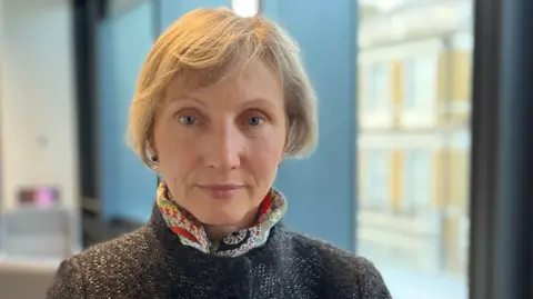 Marina Litvinenko. She has short blonde hair and is wearing a dark grey coat with a colourful, patterned scarf around her neck. She is looking directly at the camera and smiling.