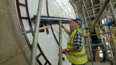Ricochet Ltd BBC Repair Shop host Steve Fletcher peers at the clock face at Manchester Town Hall, while wearing a green high-vis vest and a blue hardhat. He is stood on a scaffold with another worker. 