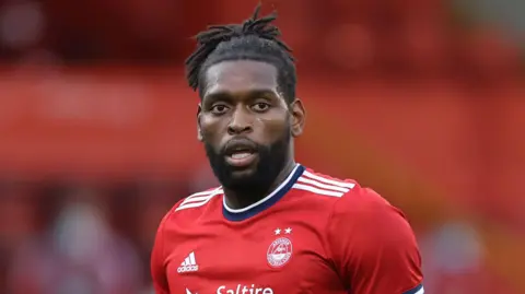 Former Arsenal winger Jay Emmanuel-Thomas looks at the camera while he is standing on football pitch. He is wearing red Arsenal uniform.
