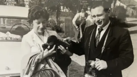 Contributed A black and white image of Victor and Denise Braybrook who are standing on a road. Denise is on the left and is holding and looking at an object. Victor is on the right and has a camera around his neck which he is holding.