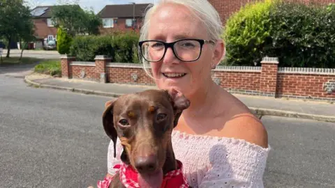 Amanda Steeles standing by the side of the road with her dog Dotty. Dotty is a brown daschund and is wearing a red collar. Amanda has a white top and black glasses