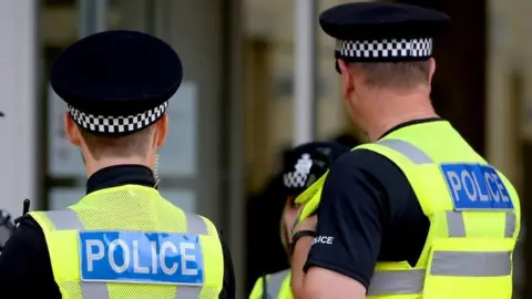 Three police officers pictured from the back wearing black polo shirts, yellow hi-vis vests with the word POLICE, and black caps with black and white chequered brims. 