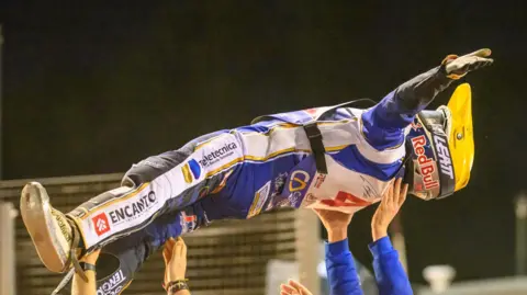 Ian Charles A speedway rider wearing a yellow helmet is held aloft on his back by people who are out of shot.