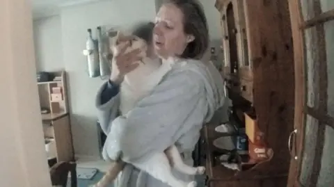 Netflix Police body-camera footage of Lucy Letby, who is wearing a grey dressing gown, tearfully holding a white and ginger cat next to a welsh-dresser style piece of wooden furniture.