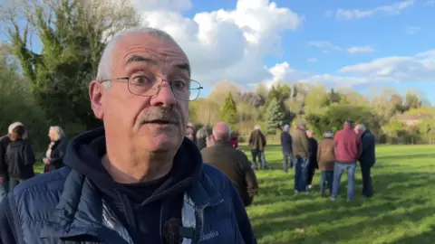 John Robertson wearing glasses and a puffer jacket and talking to the camera with crowds of people gathered behind him.