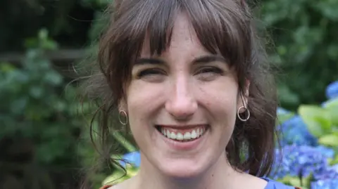 Susie Brunswick A close up picture of Susie smiling. She has tied back brown hair and is wearing two earrings.