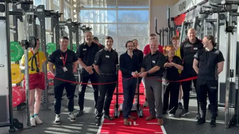 A group of 13 men and women are posing for a photograph while a red ribbon is cut to mark the opening of the gym. They are surrounded by gym weight-training machines.
