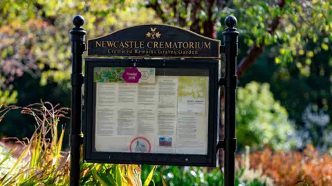 A black-framed noticeboard at Newcastle-under-Lyme Crematorium in Bradwell. There is gold lettering on a panel above the noticeboard, and there are trees and bushes in the background.
