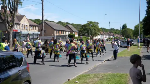 Jacob Tomlinson/BBC A group of people make their way down a street playing drums.