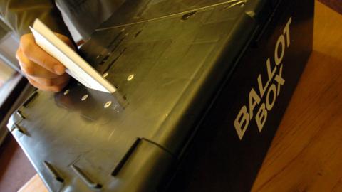 A close-up of a hand pushing a ballot paper into a black box with the words ballot box painted on the side