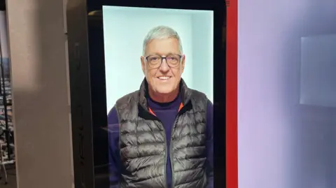 Proto Hologram Picture of hologram version of Bill on a large screen, wearing a purple jumper and black gilet and smiling.