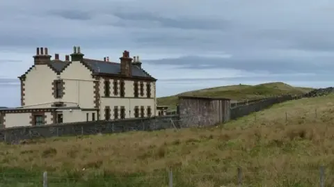 The station looks like two large houses side by side. The walls are a painted beige colour with red bricks showing along the edges of the gable ends and the windows along its walls. The station sits in an area of grassland.
