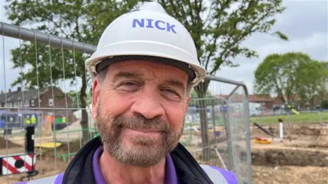 Bearded Nick Knowles smiling at the camera, wearing a white hard hat