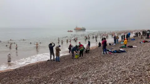 Lots of people standing on the shore of a pebble beach with many in the water in various swimming attire. It looks misty and there is an RNLI lifeboat in the distance, ahead of the crowds.