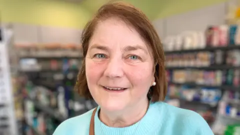 A woman with short brown hair is smiling at the camera. She is wearing a turquoise coloured jumper and is standing in front of shelves of items in a pharmacy.