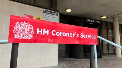 Lewis Adams/BBC A red sign that says HM Coroner's Service. It is at the entrance to Essex Coroner's Court, which is a cream building that has a black revolving door and brick paving outside.